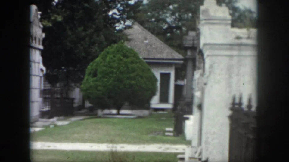 1958 New Orleans cemetery view with a clipped evergreen, white above-ground tomb, lawn, and stone gate structure.