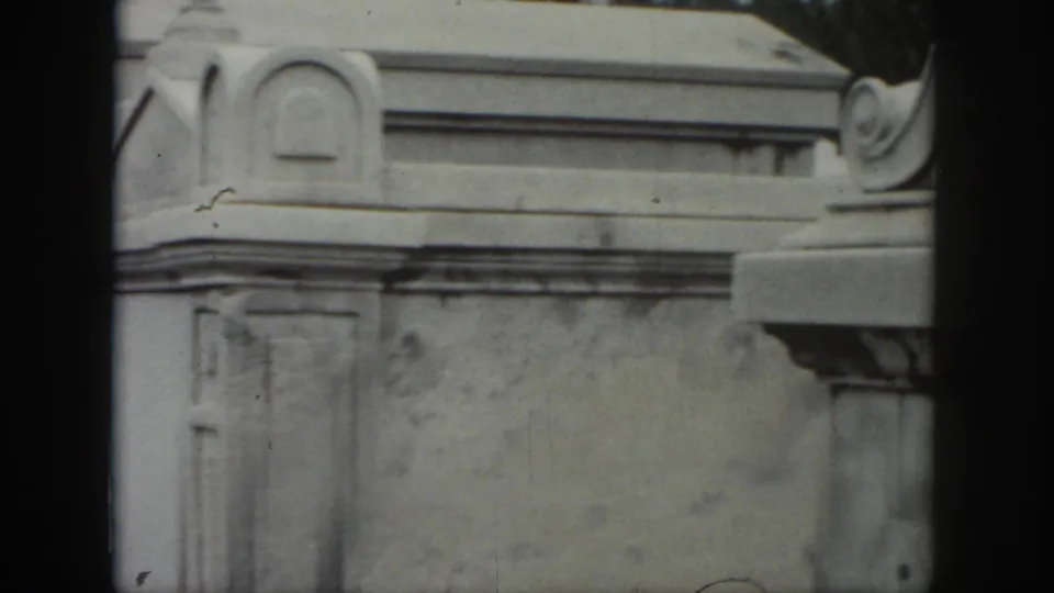 1958 New Orleans cemetery view of a pale above-ground tomb facade with carved trim, weathered stone, and partial ironwork at the edge.
