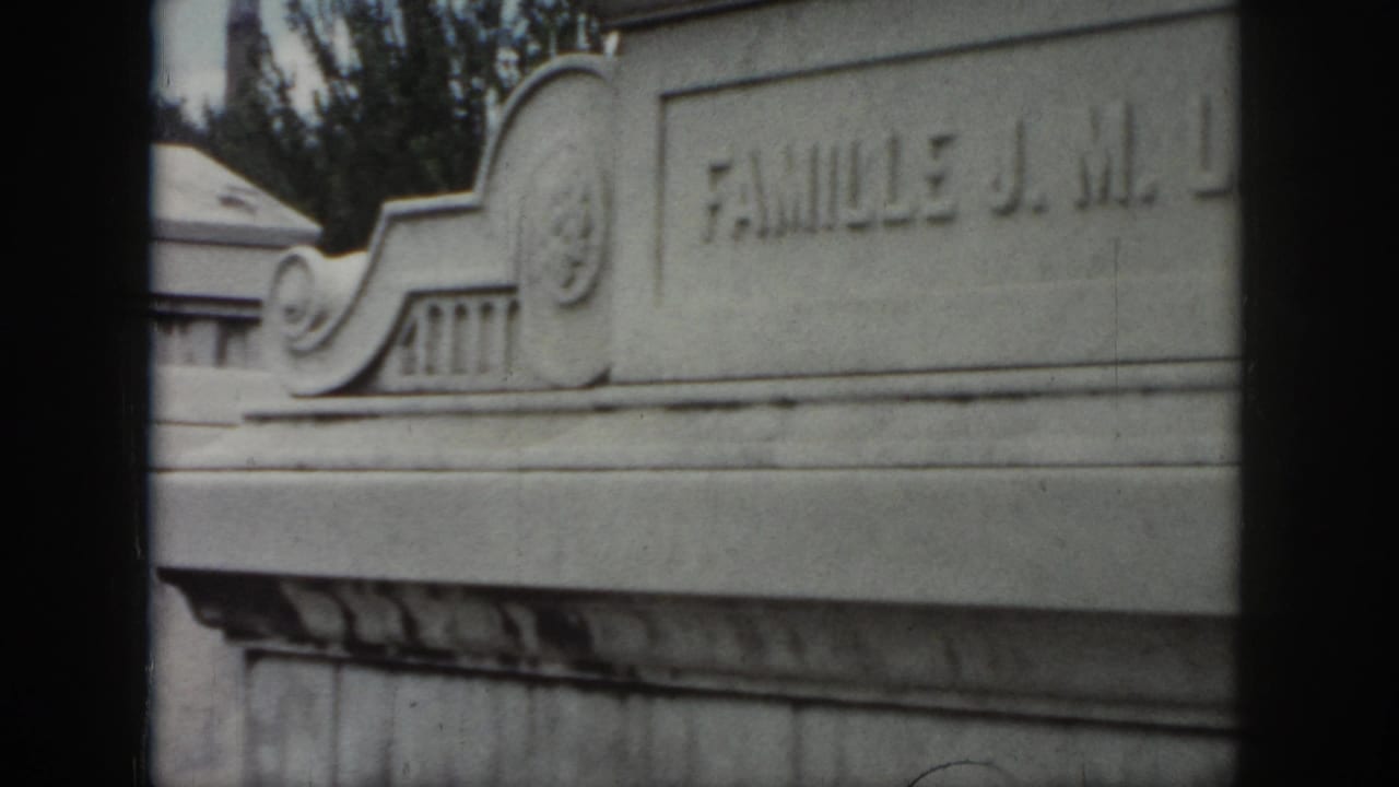 1958 New Orleans cemetery footage frame with an engraved stone tomb face, decorative top detail, and “FAMILLE J. M.” partly visible.