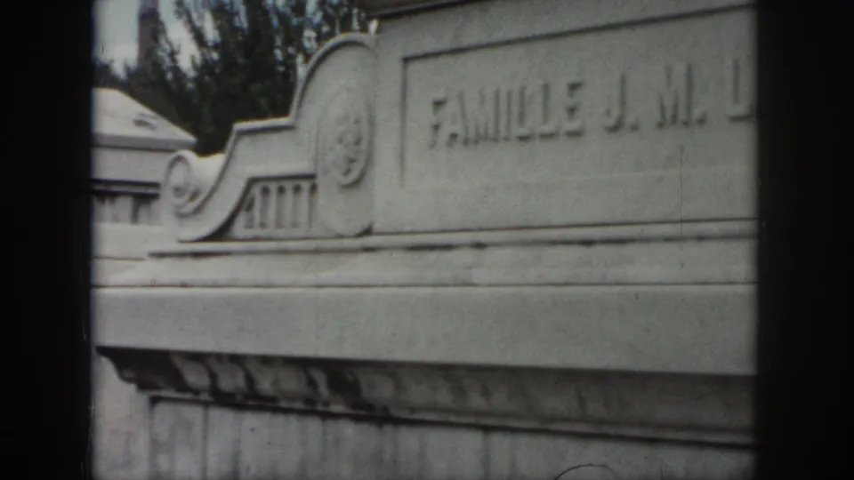 1958 New Orleans cemetery footage frame with an engraved stone tomb face, decorative top detail, and “FAMILLE J. M.” partly visible.