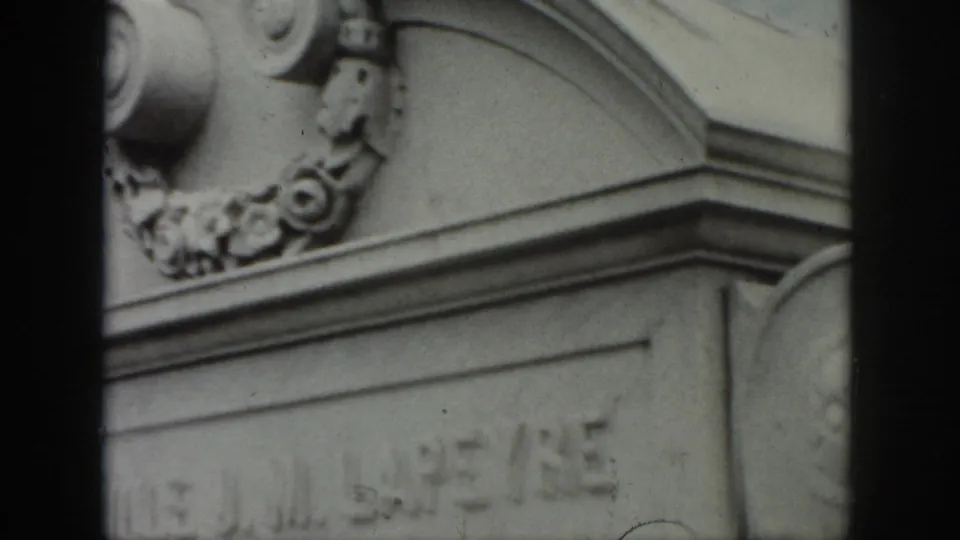 1958 New Orleans frame showing the upper portion of an ornate gray stone memorial with scroll details, floral relief, and a partly visible inscription.