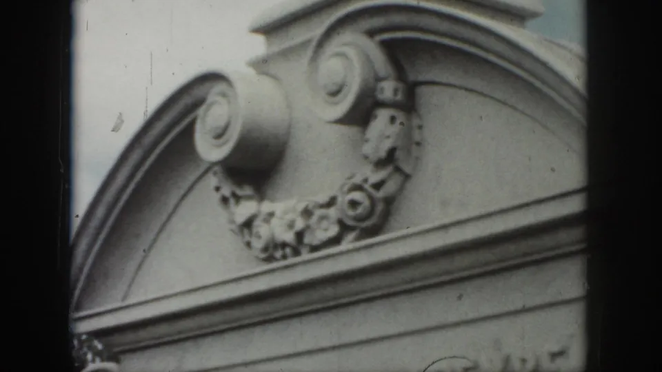 1958 New Orleans home-movie frame focused on decorative scrollwork and floral relief on a gray stone memorial above a cropped inscription.
