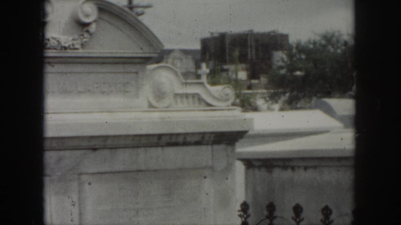 1958 New Orleans cemetery frame with a carved tomb front, decorative side scrolls, and trees beyond the monuments.