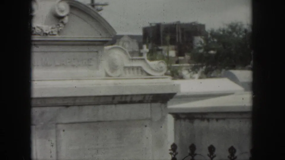 1958 New Orleans cemetery frame with a carved tomb front, decorative side scrolls, and trees beyond the monuments.
