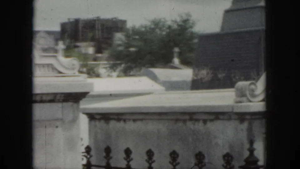 1958 New Orleans cemetery view showing above-ground tombs, iron fence finials, trees, and a darker structure behind the graves.