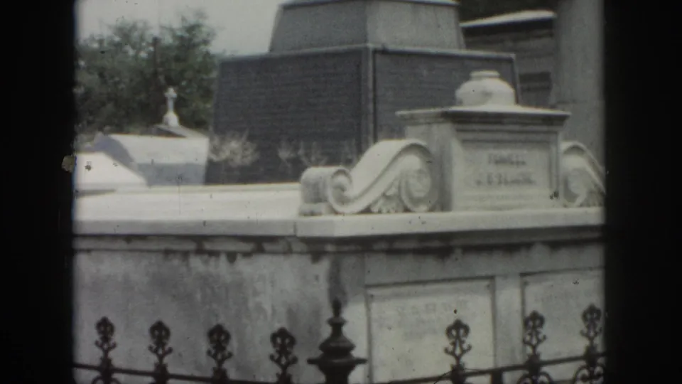 1958 New Orleans cemetery frame showing a pale stone tomb, iron fence finials, trees, and neighboring grave structures.