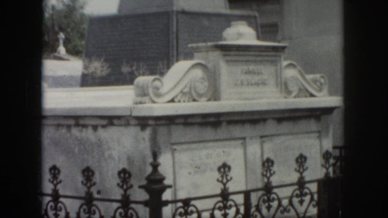 1958 New Orleans home-movie frame showing an above-ground marble tomb with carved scrollwork behind ornate iron fencing.