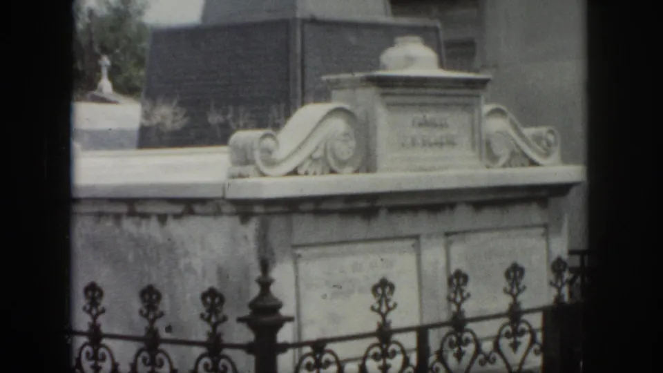 1958 New Orleans home-movie frame showing an above-ground marble tomb with carved scrollwork behind ornate iron fencing.