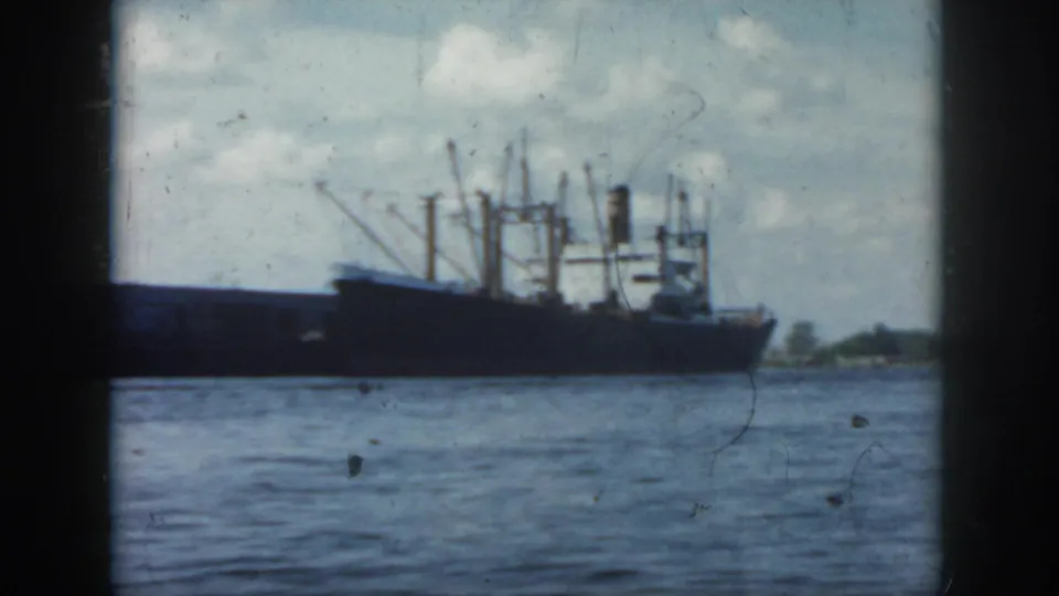 Vintage 8mm frame showing a dark cargo vessel on a New Orleans-area waterfront, with cranes or masts silhouetted against sky.
