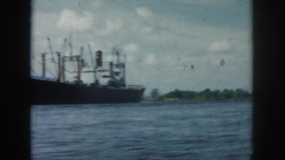 1958 home-movie frame of a dark ship on blue water, with a low distant shoreline and soft sky in the background.