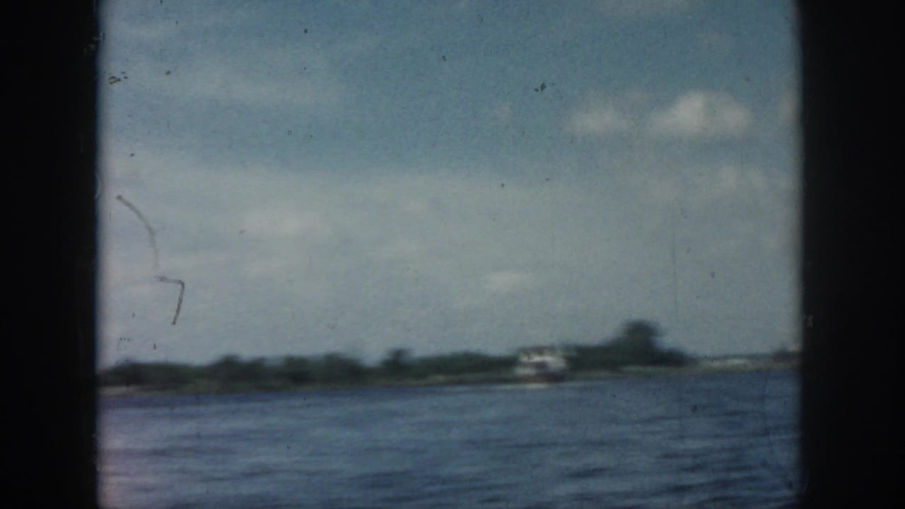 1958 New Orleans-area home-movie frame showing open water, a distant small boat, low shoreline, and partly cloudy sky.