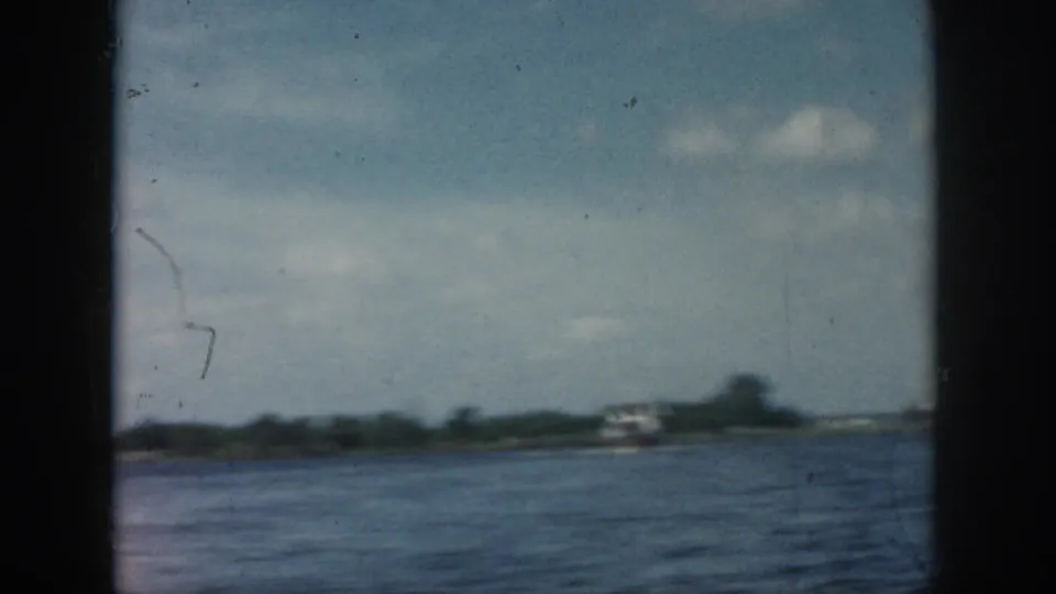 1958 New Orleans-area home-movie frame showing open water, a distant small boat, low shoreline, and partly cloudy sky.