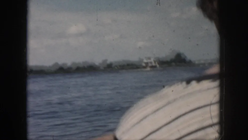 Soft 1958 home-movie frame from a moving boat, showing water, a distant small boat, shoreline, and foreground boat detail.