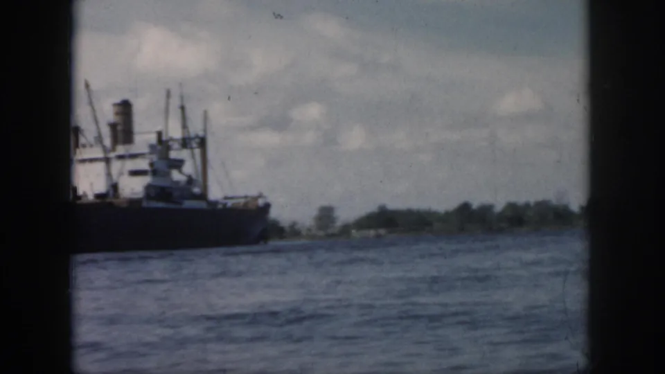 1958 New Orleans-area home-movie frame showing a large cargo ship on the water near a low shoreline under scattered clouds.