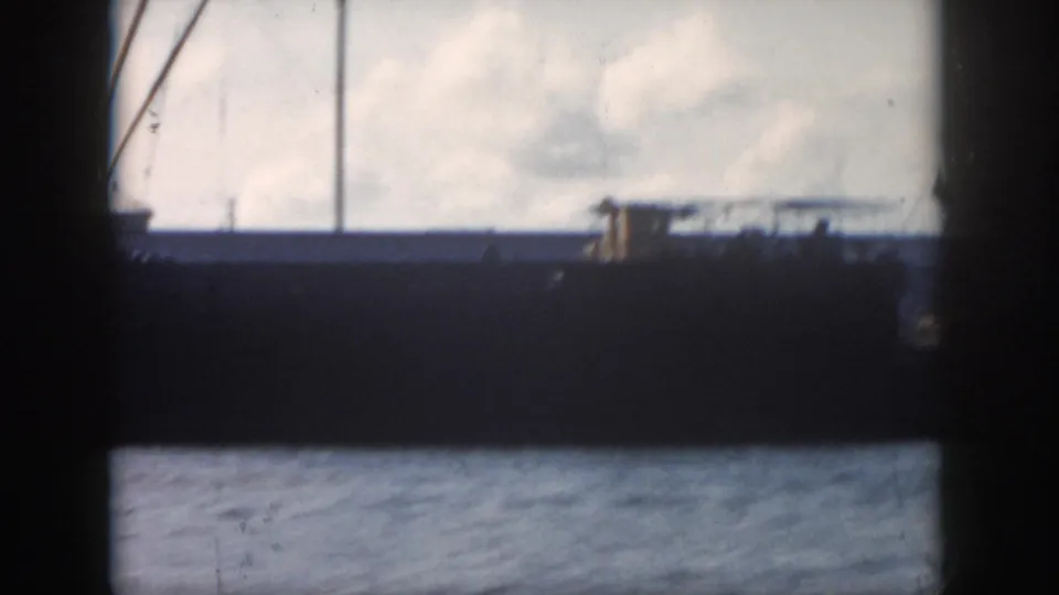 The final frame holds on the ship's dark hull, low deck details, vertical mast lines, and rippled water under a pale sky.