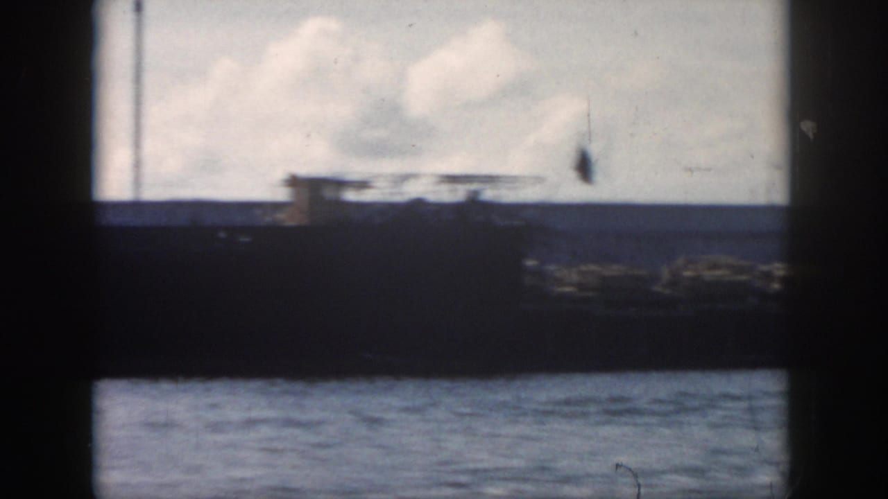 1958 New Orleans-area 8mm frame showing a large dark vessel, flags, cloudy sky, and choppy water in soft focus.