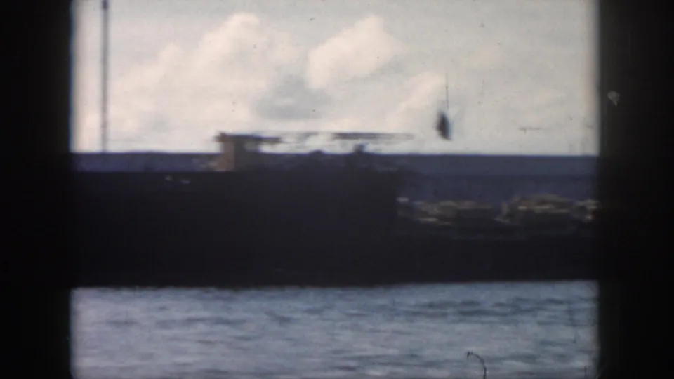 1958 New Orleans-area 8mm frame showing a large dark vessel, flags, cloudy sky, and choppy water in soft focus.