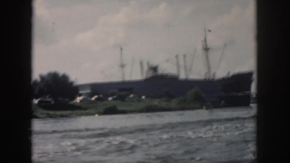 Faded 8mm frame from New Orleans showing a long ship beyond water, with trees and low vegetation along the bank.