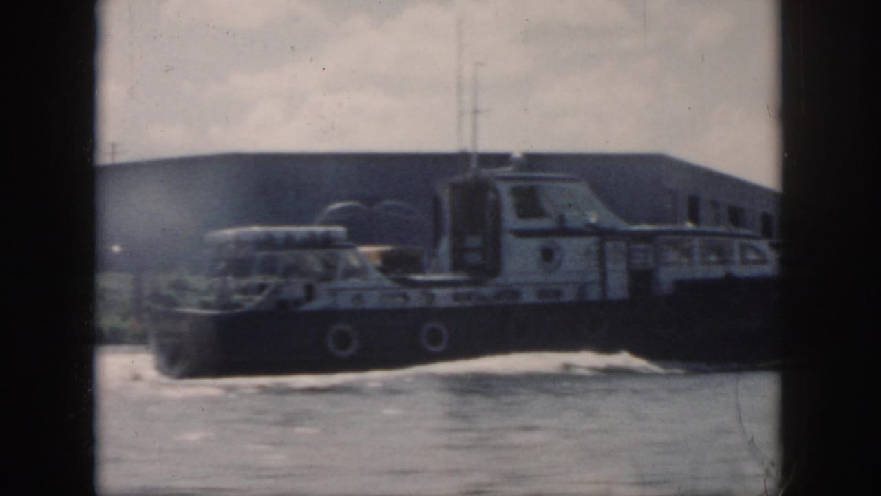 1958 New Orleans home-movie frame showing a dark cargo ship beside a long industrial dock under a cloudy sky.