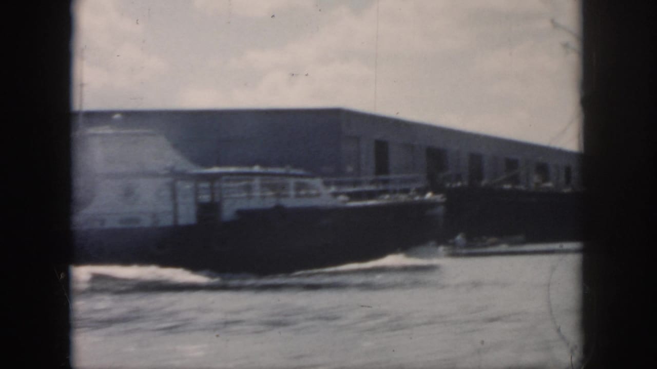 Dark cargo vessel moves across New Orleans harbor water near a long dock building in the supplied 1958 footage.