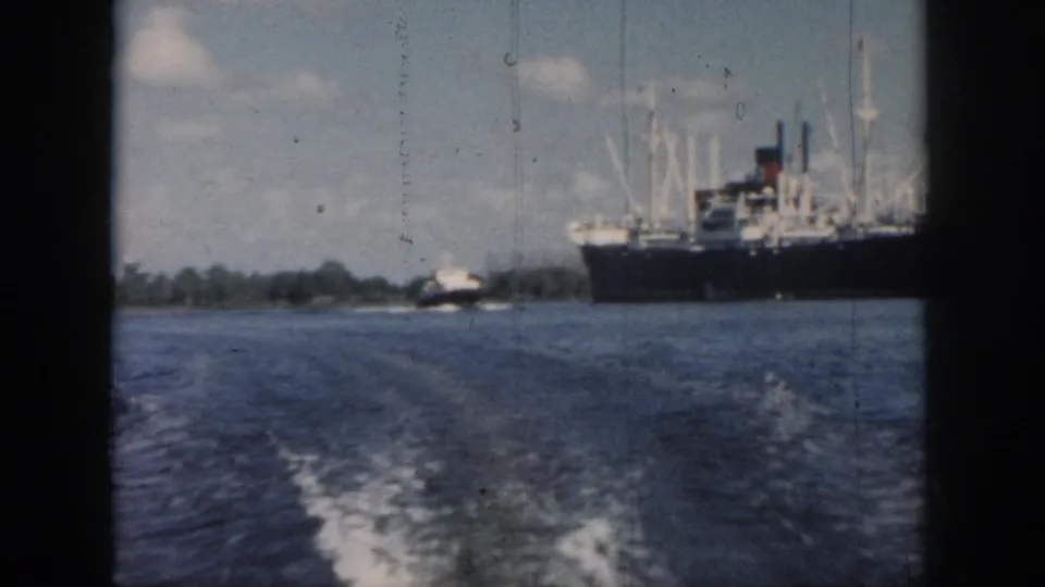 1958 New Orleans home-movie frame from a moving boat, showing wake, open water, a large ship, and a smaller vessel in the distance.