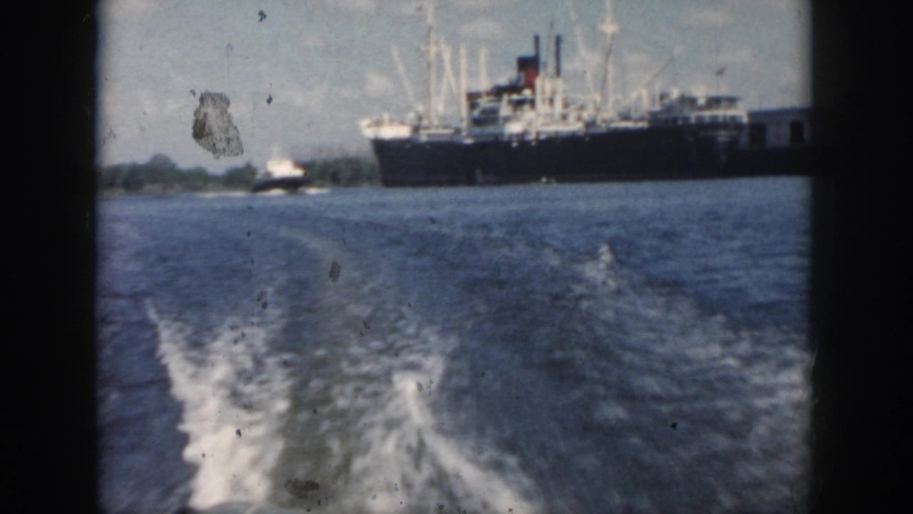 Moving-boat view in New Orleans, 1958, with foamy wake leading toward a large ship and low shoreline under a cloudy sky.