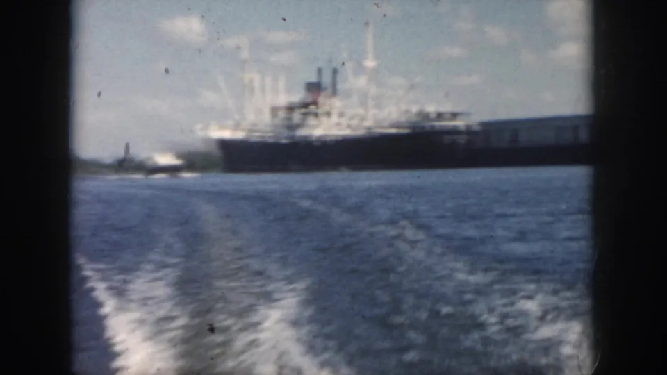 Soft 8mm frame from a moving boat in 1958 New Orleans, with a large ship stretching across the background and wake in the foreground.