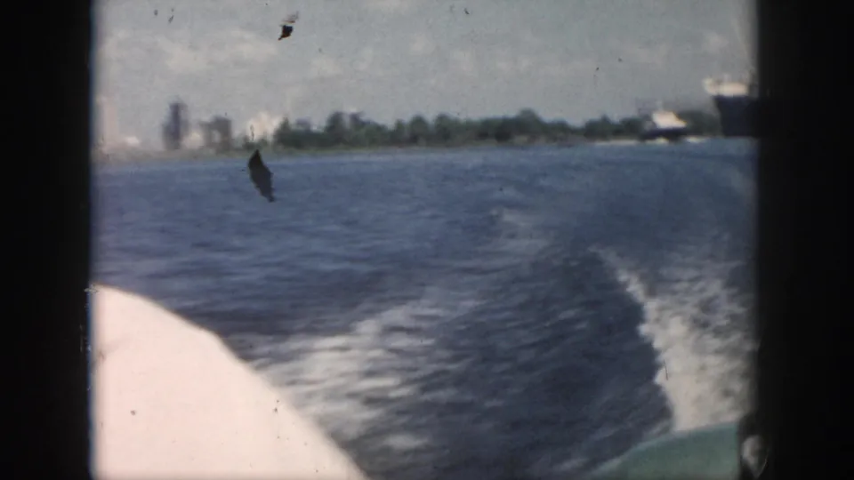 Final 1958 New Orleans boating frame shows a curving wake, open water, distant shoreline, and the edge of a large ship at right.