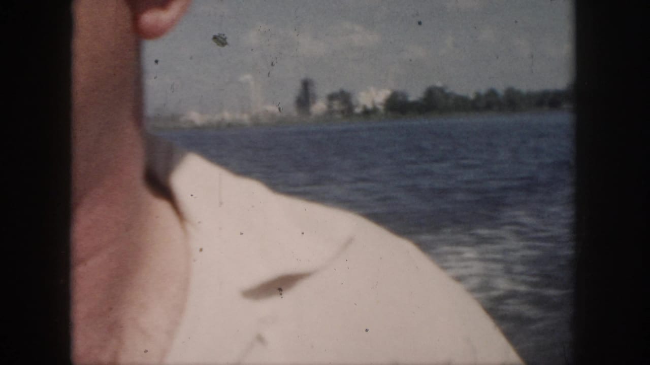 1958 New Orleans-area 8mm frame showing part of an adult man close to the camera with water and a low distant shore behind him.