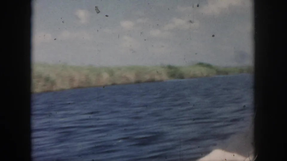1958 New Orleans-area waterway view from a boat, with rippled water, low shoreline, and cloudy daylight sky.