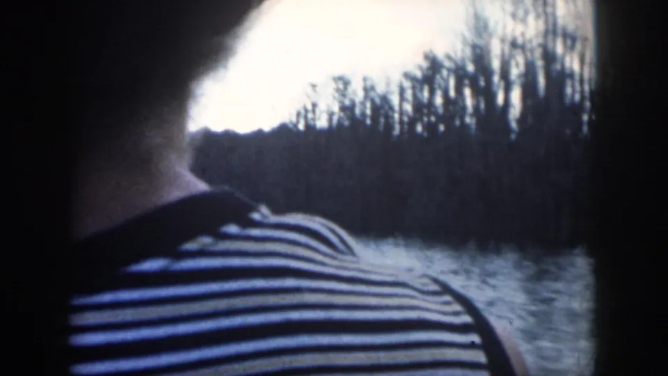 A person in a black and white striped shirt sits or stands on a boat, facing a wooded waterway in 1958 New Orleans footage.