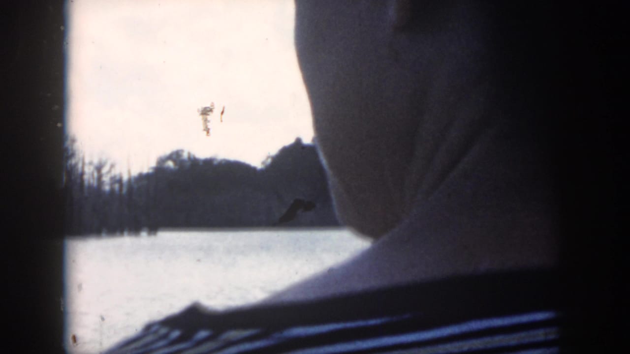 1958 New Orleans home-movie frame with a partial figure in a striped shirt silhouetted beside a window overlooking water and bare trees.