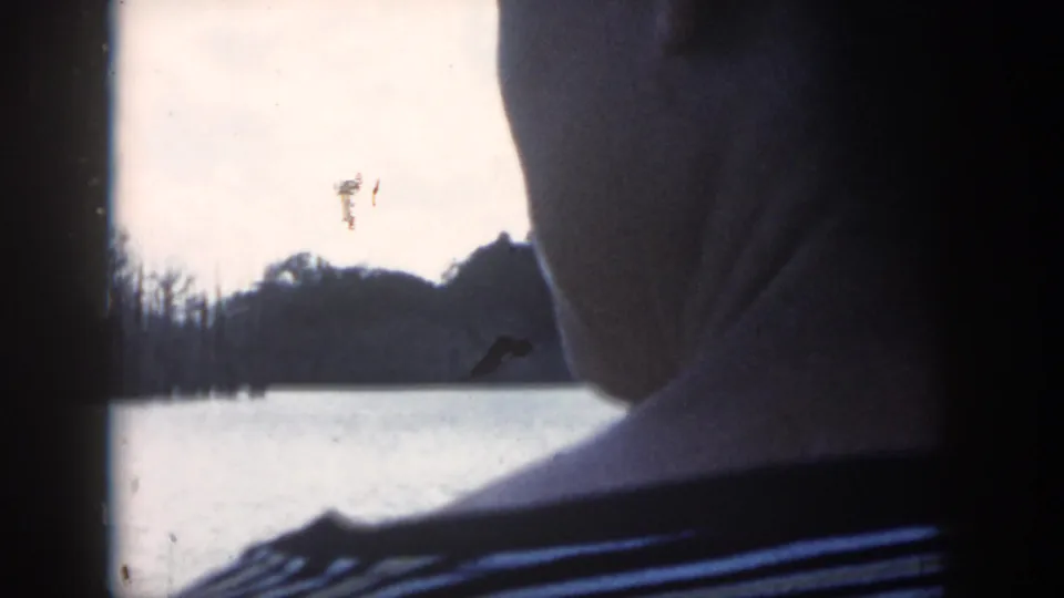 1958 New Orleans home-movie frame with a partial figure in a striped shirt silhouetted beside a window overlooking water and bare trees.