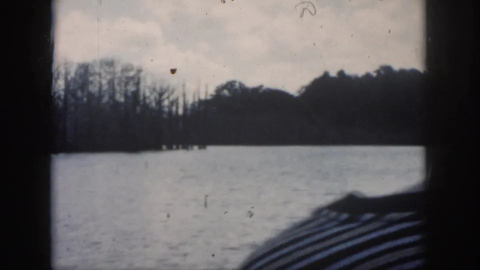 1958 New Orleans 8mm view across a calm lake through a dark window frame, with leafless trees and cloudy sky beyond.