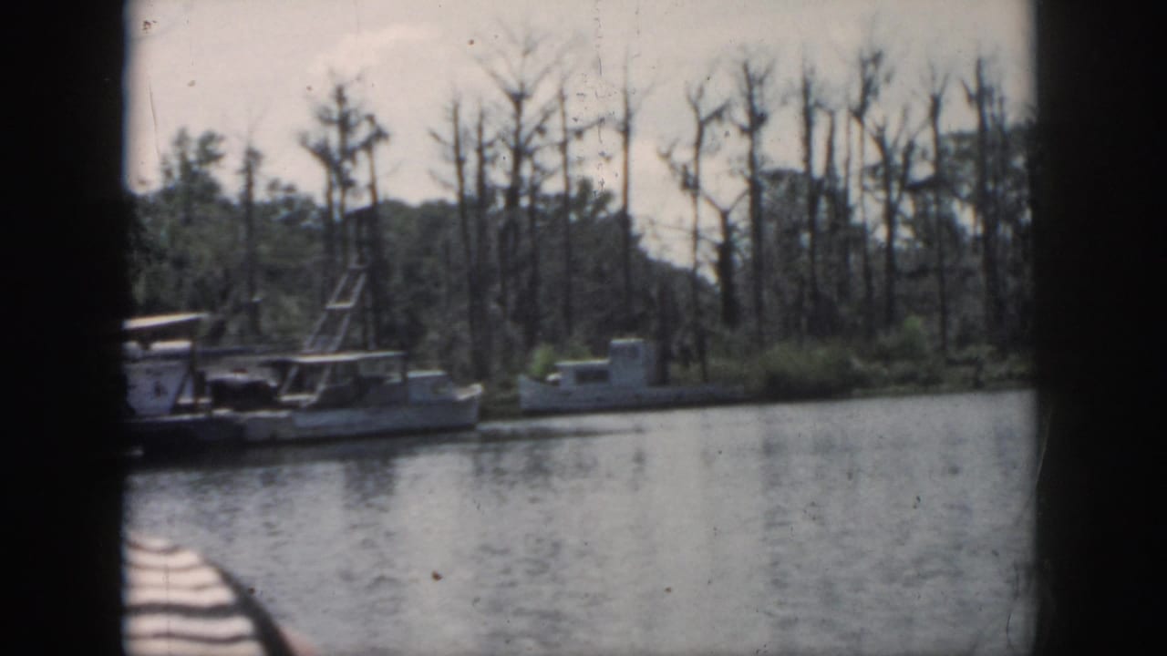1958 New Orleans-area 8mm frame showing moored small boats on calm water, with cypress trees and a dark boat edge in the foreground.
