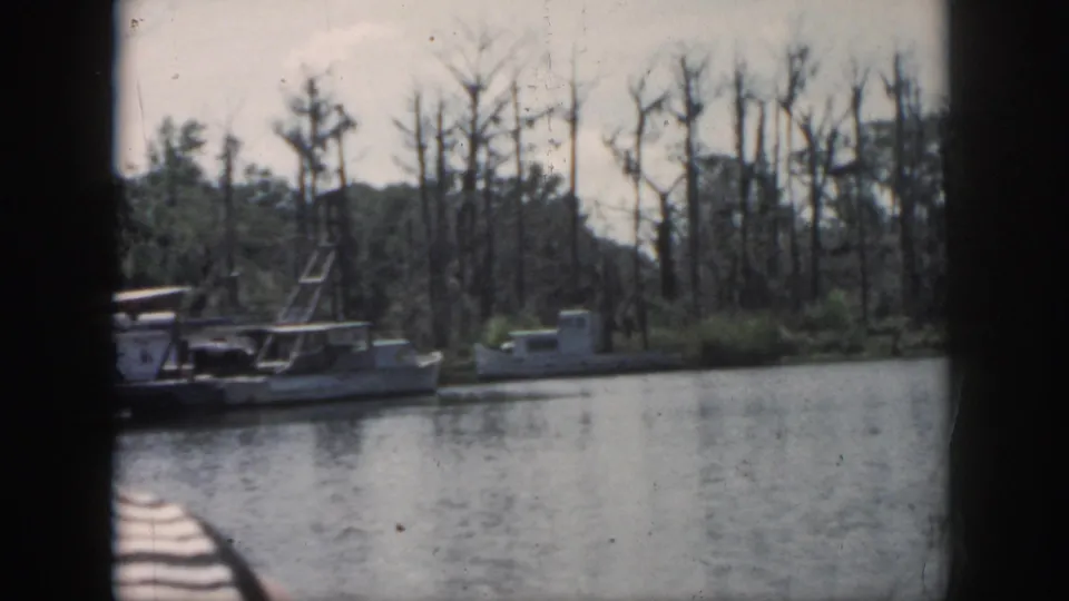 1958 New Orleans-area 8mm frame showing moored small boats on calm water, with cypress trees and a dark boat edge in the foreground.