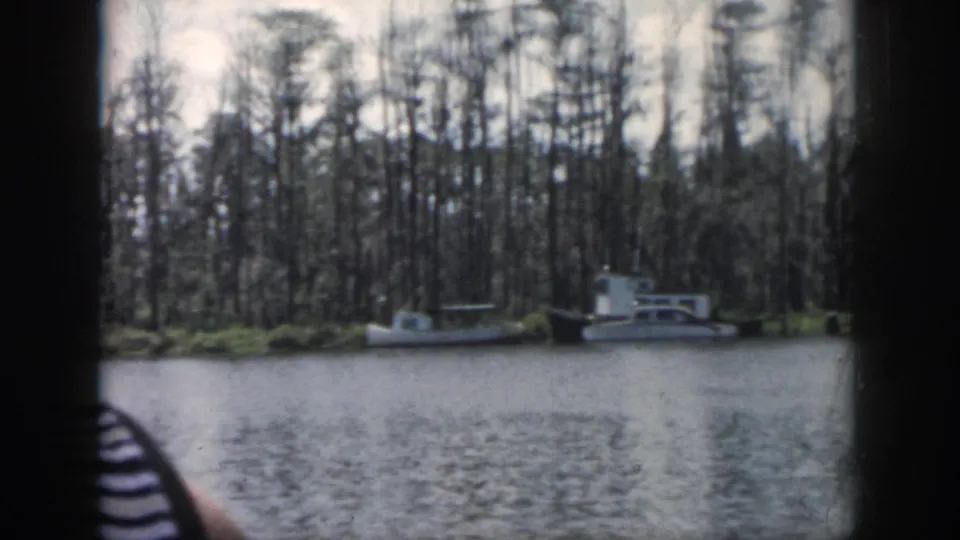 1958 New Orleans-area 8mm frame showing small boats tied near a swampy cypress bank, filmed from across rippled water.