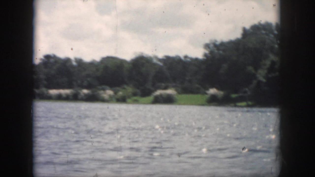 1958 New Orleans-area 8mm view across rippled river water toward trees and grass, framed by dark boat-window edges.