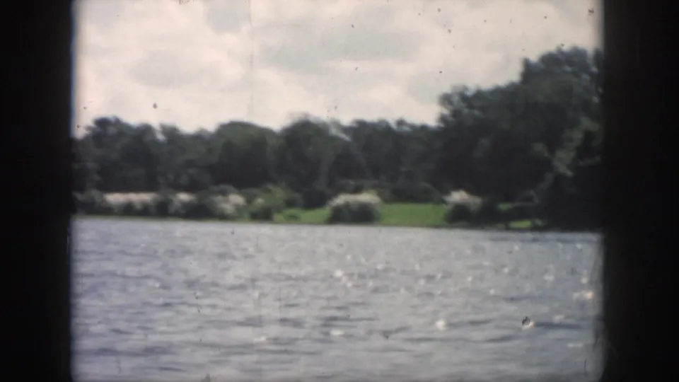 1958 New Orleans-area 8mm view across rippled river water toward trees and grass, framed by dark boat-window edges.