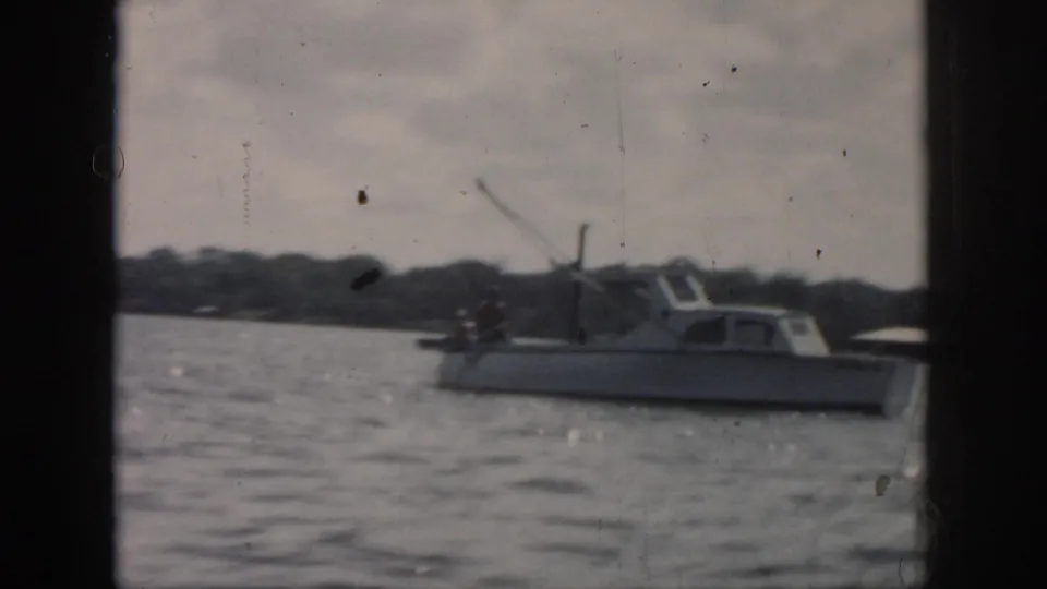 1958 New Orleans home-movie frame showing a small cabin boat on calm water, with trees and soft 8mm grain visible.