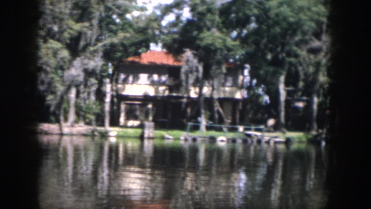 Waterside house with a red-tile roof across calm water among trees.