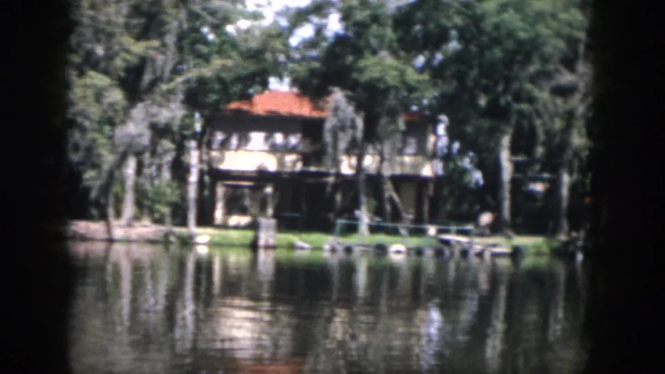 1958 New Orleans 8mm frame showing a waterside house with a red-tile roof, lawn, trees, and water reflections.