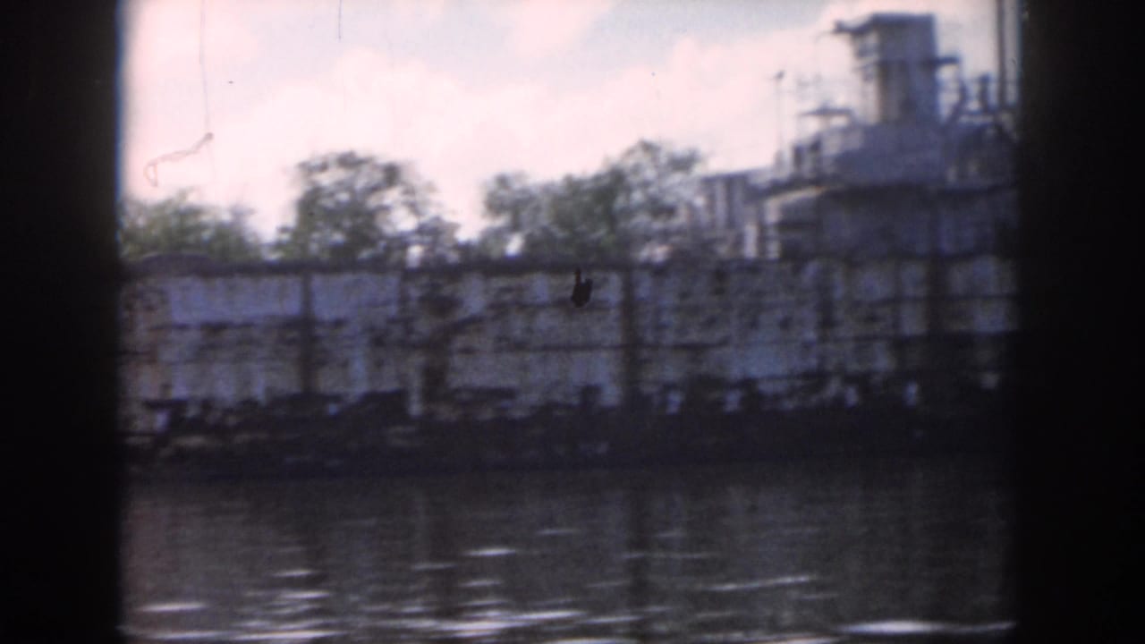 1958 New Orleans 8mm frame showing the side of a docked vessel along the water with trees and cloudy sky beyond.