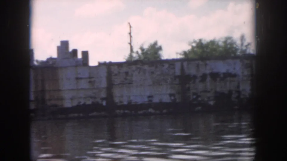 A weathered vessel and tree-lined shore in New Orleans, viewed from a moving boat through a dark vertical frame.