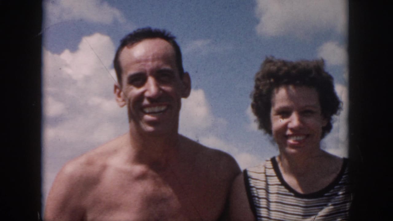 1958 New Orleans home-movie frame of a smiling couple seated close together on a boat with blue sky behind them.