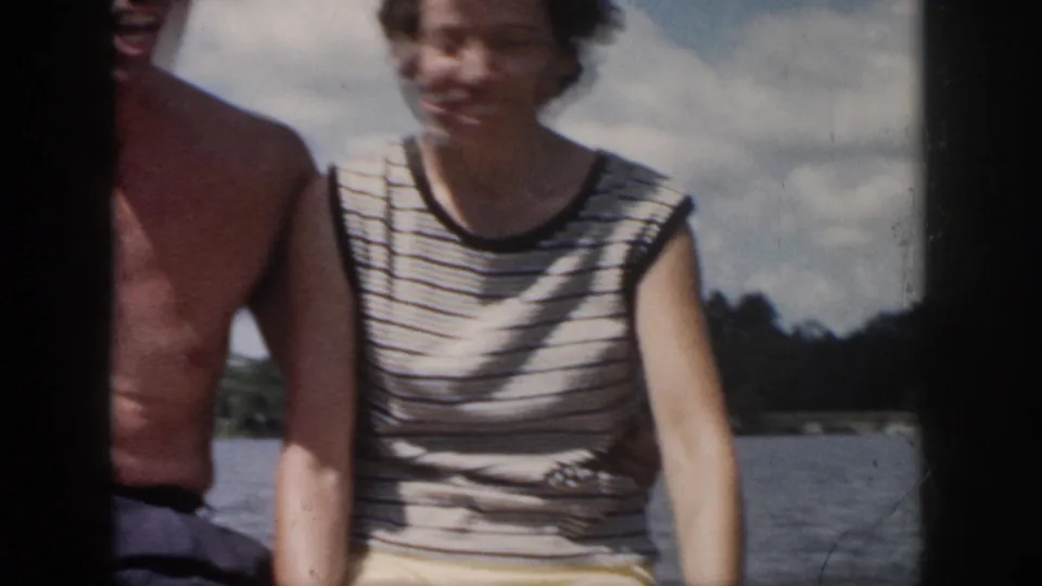 Cropped 1958 vacation frame showing the woman seated on a boat beside the man, with water and shoreline behind them.