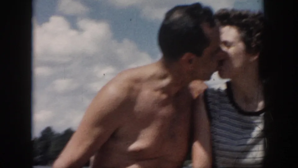 1958 New Orleans home-movie frame of the couple sharing a kiss on a boat, with water, shoreline, and clouds in the background.