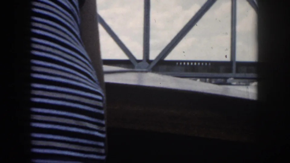 From inside a boat, a striped sleeveless top passes in front of a window framing a metal truss bridge and cloudy sky.