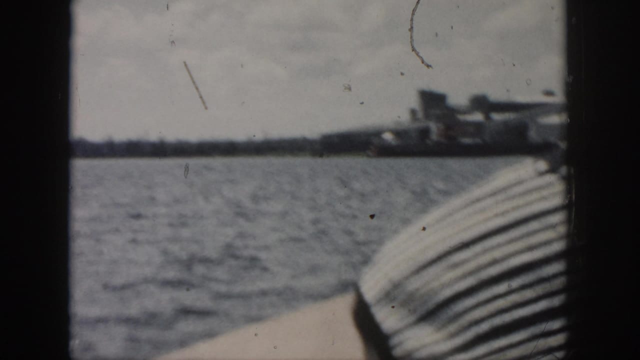 1958 New Orleans boat-view frame with rippling water, boat edge, and a soft distant industrial shoreline.