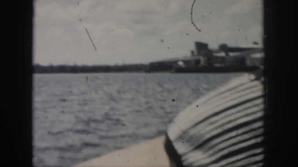 1958 New Orleans boat-view frame with rippling water, boat edge, and a soft distant industrial shoreline.
